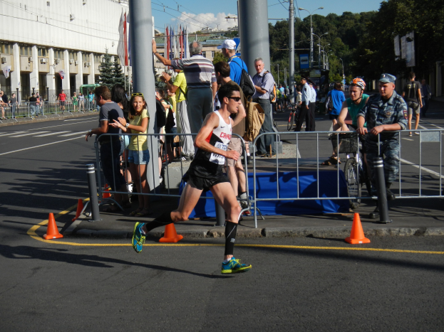 Ein Mann, der auf der Straße eines Stadtteils einen Marathon läuft, umgeben von Zuschauern und Fahrradfahrern auf dem Gehweg, unter einem bewölktem Himmel.