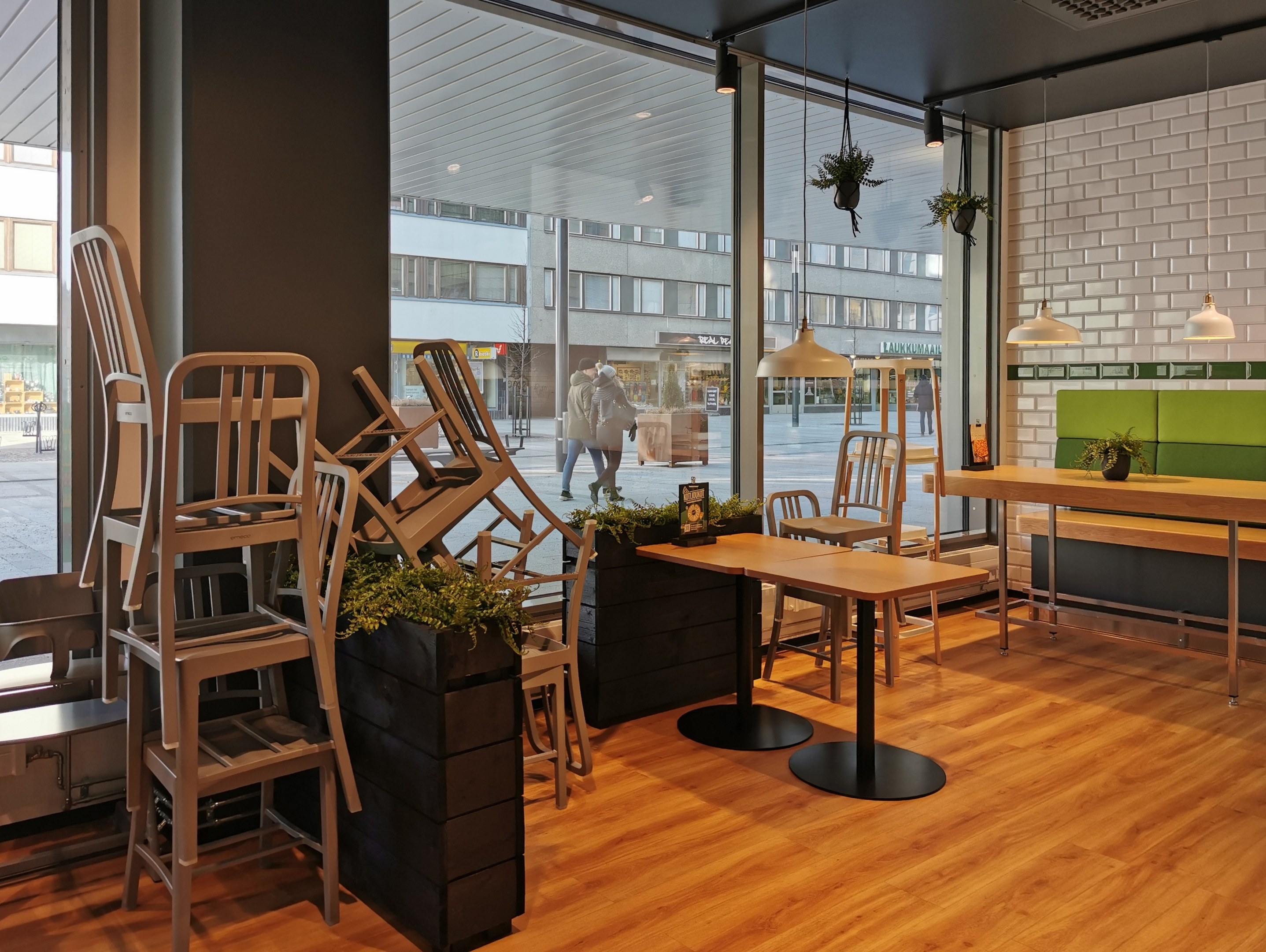 Ein Restaurantinnenraum mit hölzernen Tischen und Stühlen, sichtbar durch ein großes Fenster, das eine städtische Berliner Straßenszene mit Fußgängern, Gebäuden und urbanen Elementen zeigt.