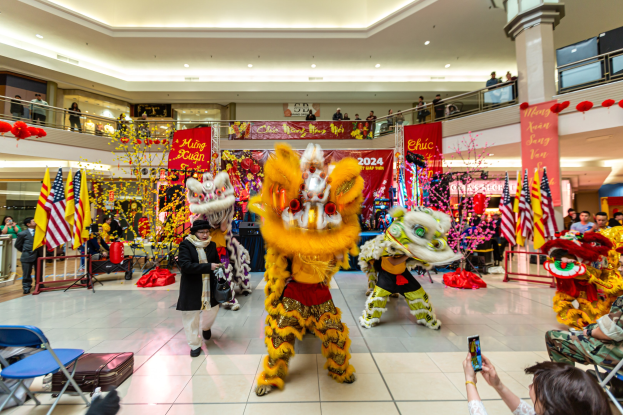 Ein lebendiges chinesisches Neujahrsfest in einem Einkaufszentrum mit Menschen in Kostümen, die tanzen, anderen, die auf Stühlen sitzen und Handys halten, sowie Flaggen, Bannern und Deckenleuchten im Hintergrund.