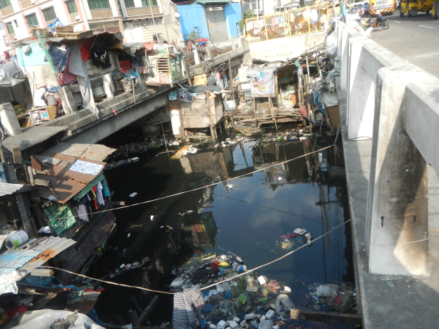 Fluss durch eine stark mit Müll verschmutzte Stadt mit einer Brücke auf der rechten Seite und Fahrzeugen auf der Straße sowie im Hintergrund sichtbare Gebäude.