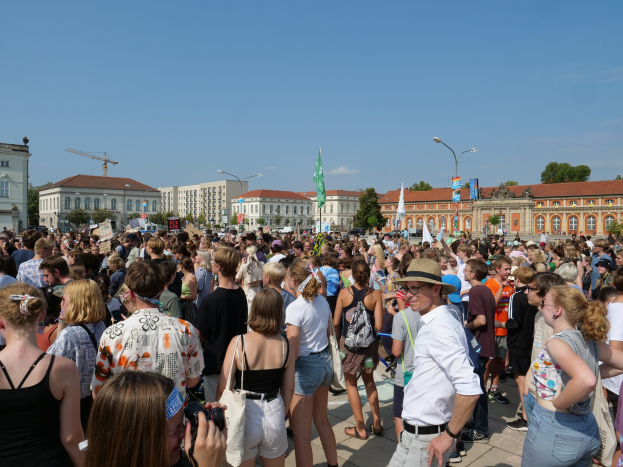 Large crowd of people gathered outside a building with flags, trees, and light poles in the background, some holding cameras during a protest or demonstration.