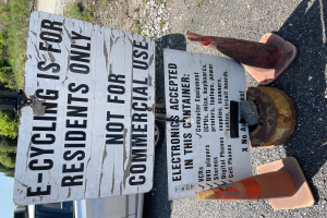 Ein Schild mit der Aufschrift "E-Bike-Fahren ist nur für Bewohner" steht an der Seite einer Straße, umgeben von Verkehrskegeln und anderen Gegenständen, mit einem Fahrzeug, Bäumen und einem klaren blauen Himmel im Hintergrund.