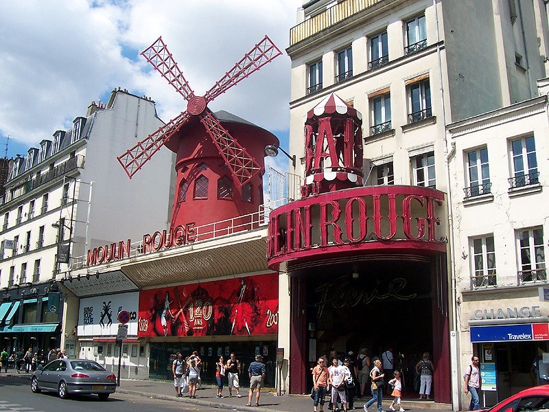 Eine belebte Straßenszene vor dem Moulin Rouge in Paris mit Passanten, Fahrzeugen, Gebäuden und einer Windmühle im Hintergrund unter einem bewölkten Himmel.
