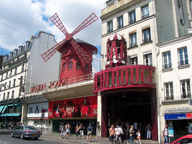 Eine belebte Straßenszene vor dem Moulin Rouge in Paris mit Passanten, Fahrzeugen, Gebäuden und einer Windmühle im Hintergrund unter einem bewölkten Himmel.