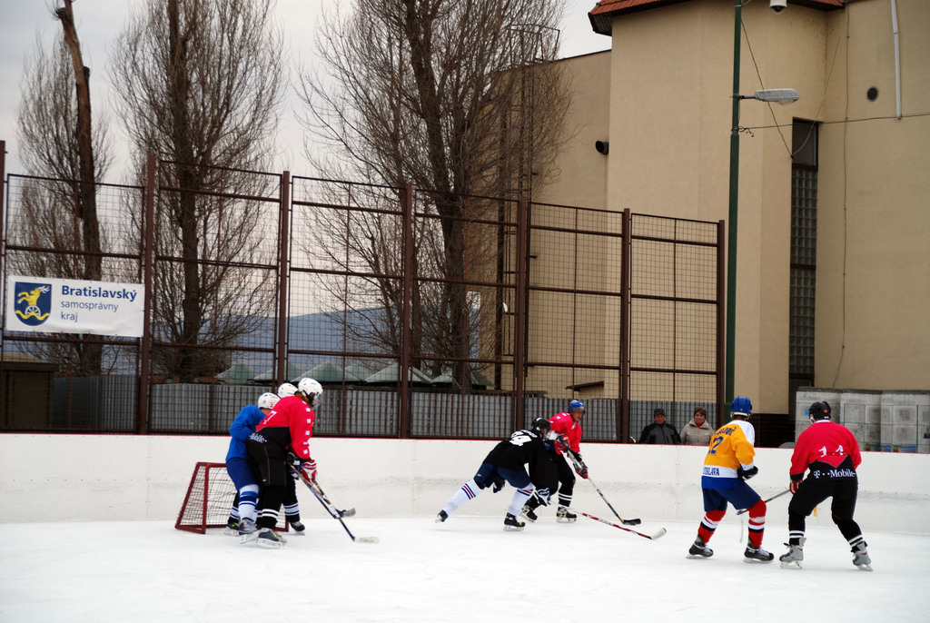 Personen spielen Eis Hockey auf einer Eisfläche mit Gebäuden, Bäumen, einer Straßenlaterne, einer Namensplakette und Zäunen im Hintergrund unter einem Himmel.