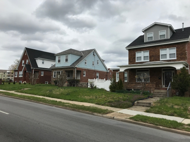 Eine Reihe von Häusern an einer Straße mit sichtbaren Renovierungswerkzeugen und -materialien, umgeben von Gras, Pflanzen, Bäumen und einem bewölkten Himmel.