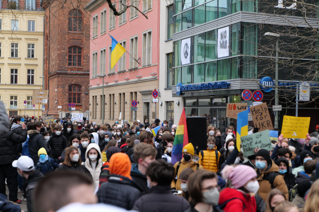 Eine große Gruppe von maskierten Menschen mit Schildern protestiert gegen ein Regierungsverbot der Maskenbenutzung in einer Stadtstraße, umgeben von Gebäuden, Schildern, Flaggen und Bäumen.
