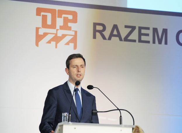 Ein Mann in Anzug und Krawatte steht an einem Podium mit einem Glas Wasser und zwei Mikrofonen und spricht zu einem Event mit einem "Razem Global"-Banner dahinter.