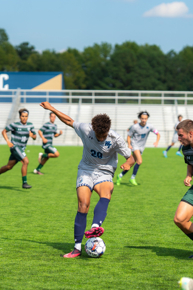 Eine Gruppe junger Männer, die Fußball auf einem Feld mit Bäumen und einem klaren blauen Himmel spielen, wobei ein Spieler Turnschuhe trägt.