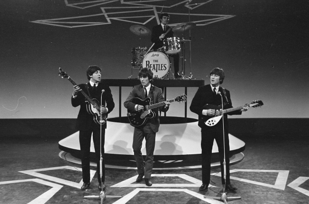 The Beatles perform on stage at the BBC Studios in London, with three members holding guitars and singing into microphones while a drummer plays behind them in a black-and-white image.