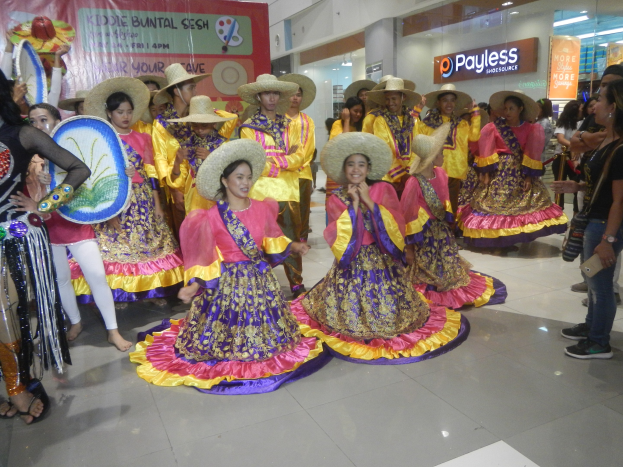 Eine Gruppe von Menschen in bunten Kleidern und Hüten, die in einem Einkaufszentrum tanzen, mit anderen, die in der Nähe stehen und Musikinstrumente sowie ein Banner mit Text halten, vor einem festlichen Hintergrund mit Schildern, Deckenleuchten und Dekorationen.