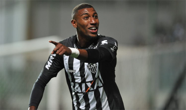 Ein Fußballspieler in einem schwarz-weißen gestreiften Trikot feiert mit einem Lächeln nach einem Tor, der Hintergrund unscharf, um den Moment zu betonen.