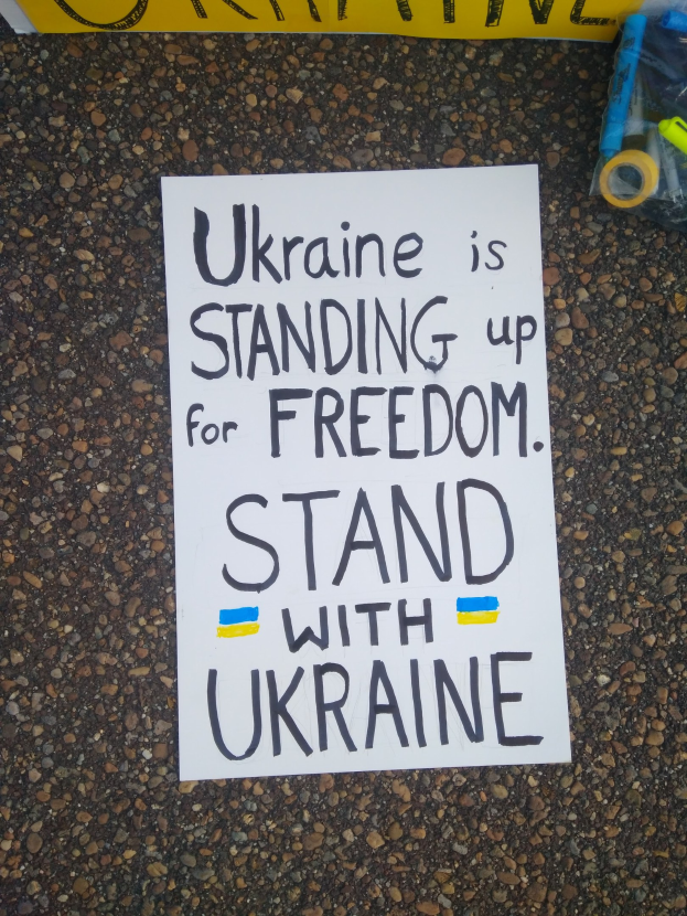 Weißer Zettel mit 'Ukraine steht für Freiheit, steh mit Ukraine' Text auf dem Boden vor einer gelben Fahne, mit einer Plastikhülle mit verschiedenen Gegenständen rechts.