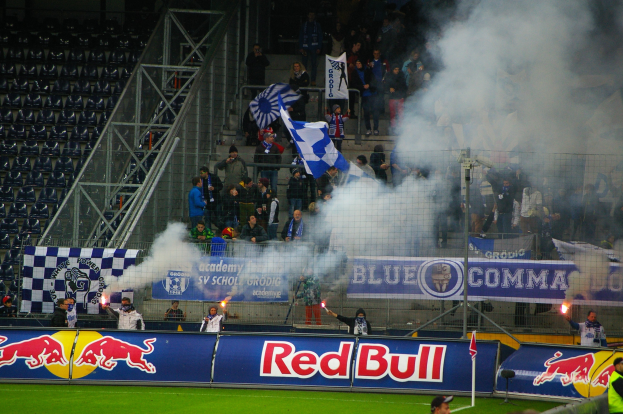 Eine Gruppe von Menschen auf einem Fußballfeld, die Fahnen und Schilder halten, mit Rauch, der aus dem Boden aufsteigt, umgeben von einem Metallzaun und leeren Stadionstühlen.