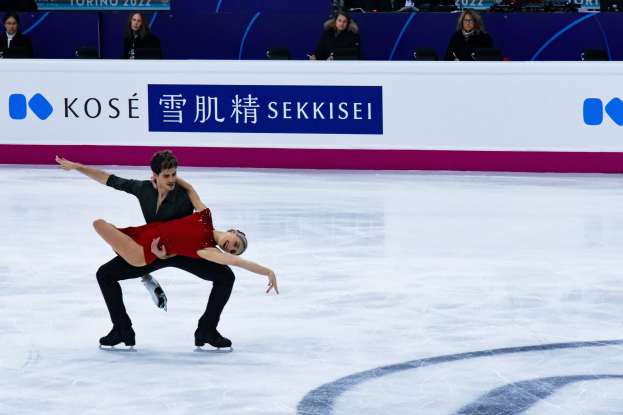 Zwei Eiskunstläufer gleiten synchron auf einer Eisbahn während eines Wettbewerbs, mit einem Publikum und einer Anzeigetafel im Hintergrund.