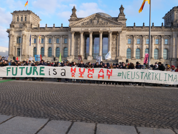 Group of people holding a banner reading "Future is a Human" in front of the Reichstag building in Berlin, Germany, with the building's architectural details visible and flags in the background under a cloudy sky.