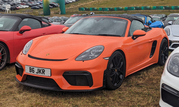 Roter Porsche 718 Boxster GTS auf dem Goodwood Festival of Speed mit Autos, einem Banner, Bäumen und einem klaren blauen Himmel im Hintergrund.