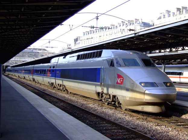 Hochgeschwindigkeitszug auf Schienen neben einem Bahnsteig mit Gebäuden und einem klaren blauen Himmel im Hintergrund.