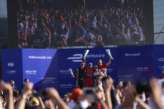 Gruppe von Menschen auf einem Formel E-Podium in Mexiko-Stadt, die Pokale halten, mit Werbetafeln und einem Bildschirm im Hintergrund.