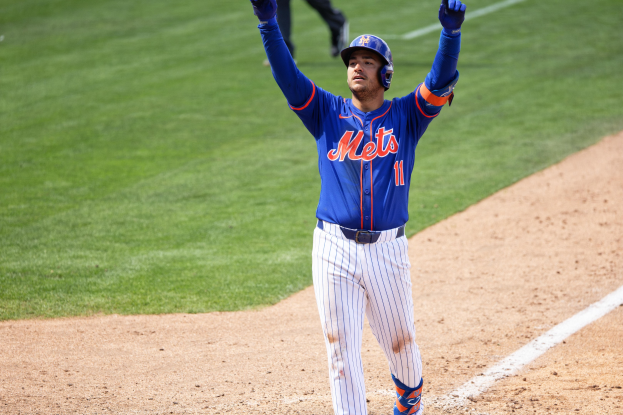 Ein Baseballspieler in einem blauen T-Shirt, weißen Hosen, einem blauen Helm und blauen Handschuhen steht auf einem grünen Feld und feiert mit den Beinen eines anderen Menschen im Hintergrund.