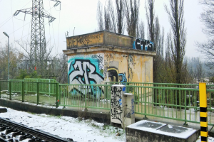 Ein Bahngeleise mit Graffiti an der Seite, umgeben von Geländern, Pfählen, Lichtern und Bäumen, mit Türmen und Drähten im Hintergrund und Schnee am Boden.