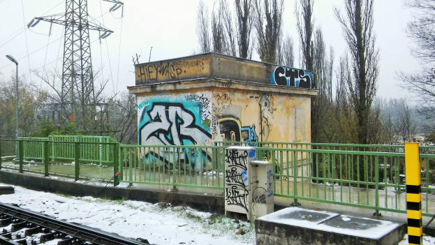 Ein Bahngeleise mit Graffiti an der Seite, umgeben von Geländern, Pfählen, Lichtern und Bäumen, mit Türmen und Drähten im Hintergrund und Schnee am Boden.