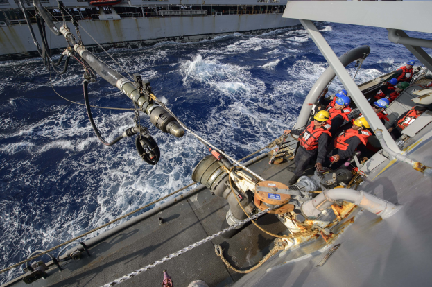 Gruppe von Menschen in Lebenswesten und Helmen auf einem Boot im Meer mit einem Schiff im Hintergrund unter einem klaren blauen Himmel, Seile und Rohre sind auf dem Boot sichtbar.