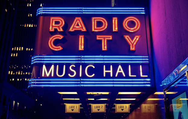Hell erleuchtetes Radio City Music Hall in New York City mit hohen Gebäuden im Hintergrund, das eine 'Radio City'-Leuchtschrift und mehrere kleinere Schilder auf der rechten Seite zeigt.