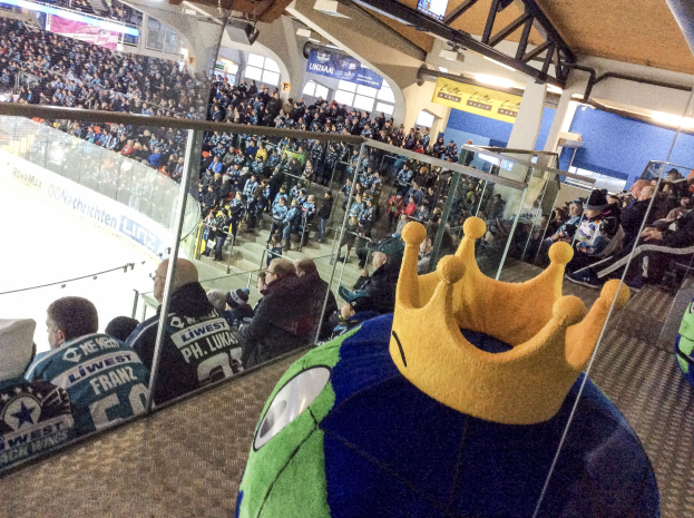 Menge im Stadion bei einem Hockey-Spiel, mit einer Person mit Krone im Vordergrund, einer Glaswand dahinter, Bannern, Säulen und Deckenleuchten.