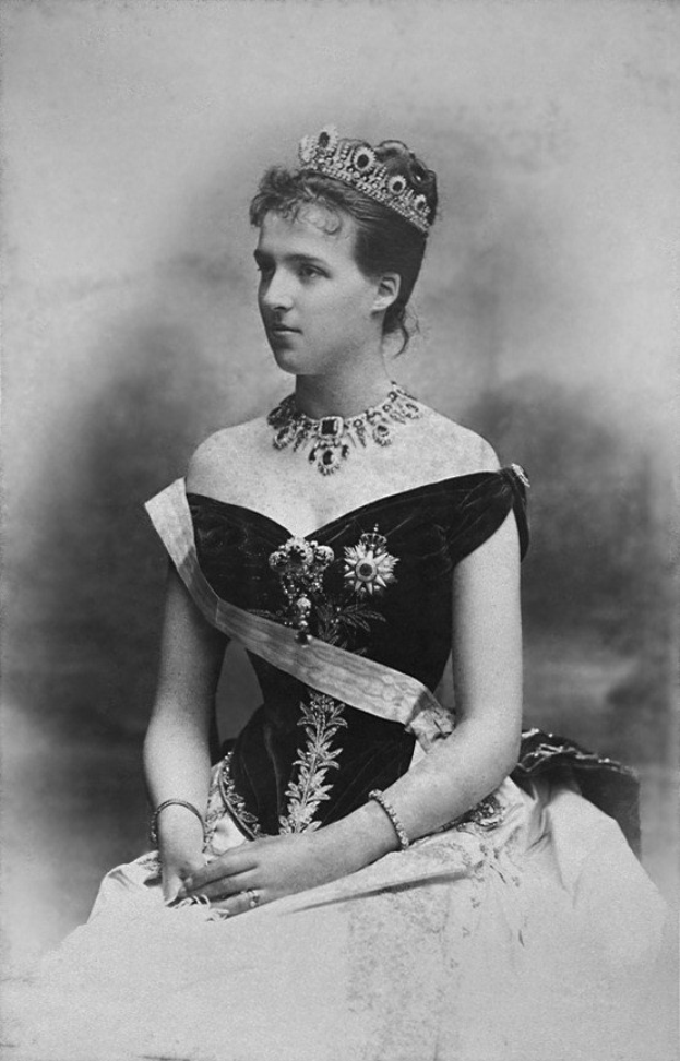 Ein Schwarz-Weiß-Foto einer Frau in einem hochgeschlossenen, langärmeligen Kleid mit einer Tiara, deren Haar elegant hochgesteckt ist.