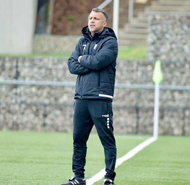 A man in a black jacket and pants stands on a soccer field with arms crossed, appearing to be a coach during a training session, with a fence, stairs, and a wall in the background.