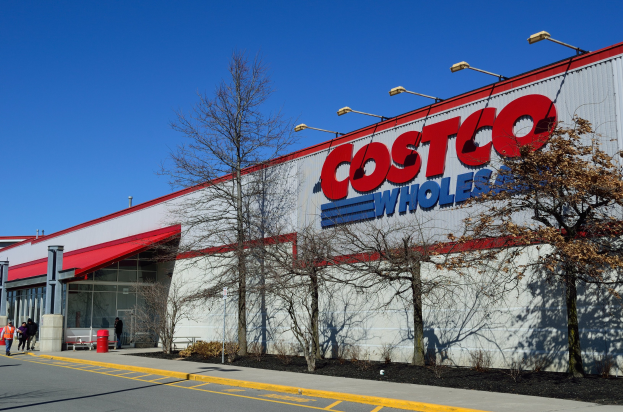 Costco Wholesale-Laden mit rotem Vordach, umgeben von Bäumen, Pfählen, Lampen und einer Straße, mit ein paar Menschen vor dem Gebäude und dem Himmel im Hintergrund.