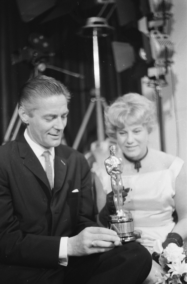 Ein Mann in einem schwarzen Blazer, weißem Hemd und Krawatte hält eine Oscar-Statue in der Hand und sitzt neben einer Frau, die einen Blumenstrauß hält, beide in eleganter Kleidung, mit abgedunkeltem Hintergrund in Schwarz-Weiß.