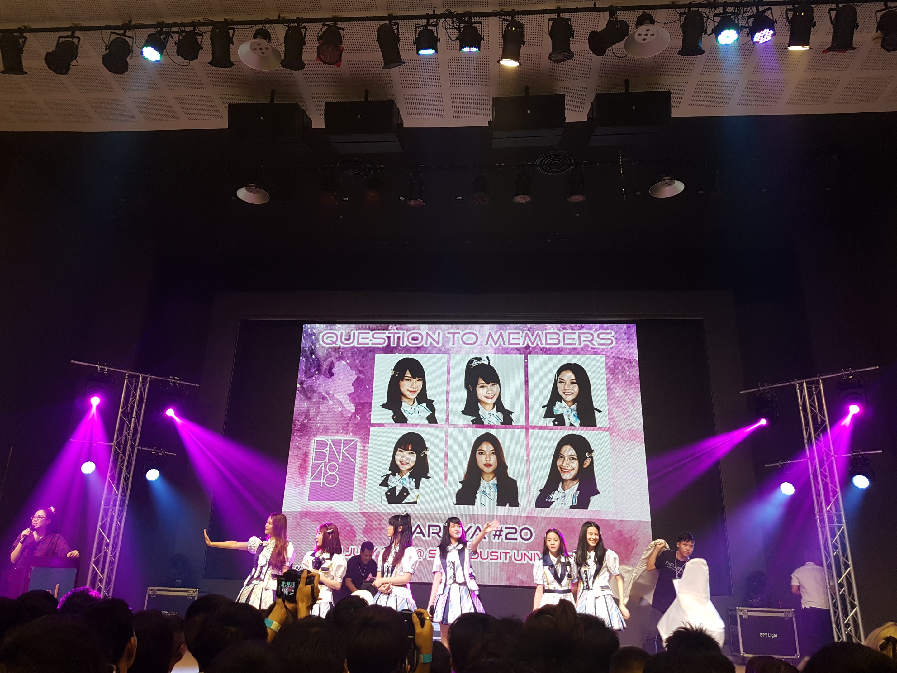 A group of people performing on a brightly lit stage with a crowd below, featuring a person holding a microphone and another playing an instrument, while a large screen displays images of BK48.