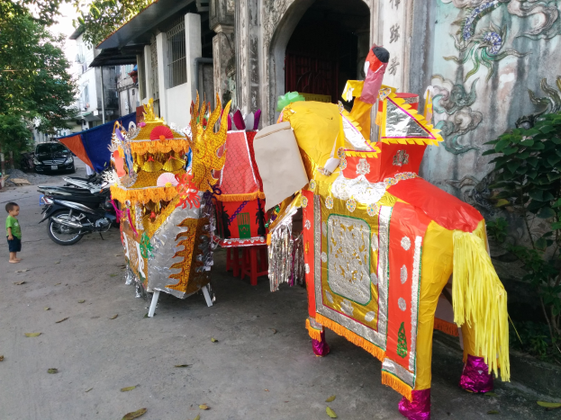 A vibrant Chinese New Year parade in Ho Chi Minh City, Vietnam, with vehicles, a person on the left, buildings and trees in the background, a painted wall on the right, and decorative parade items in the foreground.