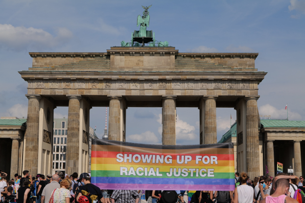 Eine Gruppe von Menschen steht vor dem Brandenburger Tor in Berlin, Deutschland, und hält eine "Racial Justice"-Schriftrolle in den Händen, mit den Säulen des Tors, einer Statue, Gebäuden und einem bewölkten Himmel im Hintergrund.