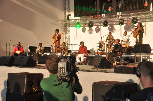 Ein musikalisches Konzert auf der Bühne mit S├Ąngern und Musikern, die Gitarre und Schlagzeug spielen, beleuchtet von roten und grünen Lichtern, mit Lautsprechern unten und zwei Personen - eine Aufnahme in einem grünen Hemd und eine andere, die zuh├Ârt.