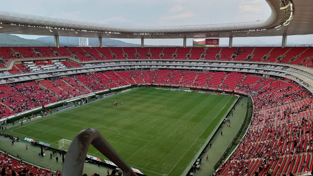 Großes Stadion voller Zuschauer bei einem Fußballspiel, mit sitzenden Fans in den Rängen, einem Geländer unten, Hügel im Hintergrund und einem klaren blauen Himmel.