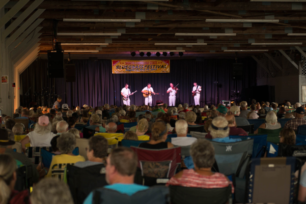 Eine große Zuschauermenge sitzt einer Bühne gegenüber, auf der Musiker mit Instrumenten spielen, Lautsprecher und Foto-Rahmen im Veranstaltungsort sichtbar sind.