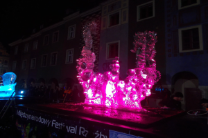 Eine Gruppe von Menschen versammelte sich um eine beleuchtete Eisskulptur auf einer nächtlichen Straße, mit einem Schild im Vordergrund und Gebäuden im Hintergrund.