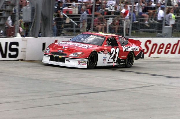 NASCAR-Auto auf einer Rennstrecke mit Zuschauern hinter Maschendrahtzaun, einige sitzen und einige stehen, mit Schildern im Hintergrund.