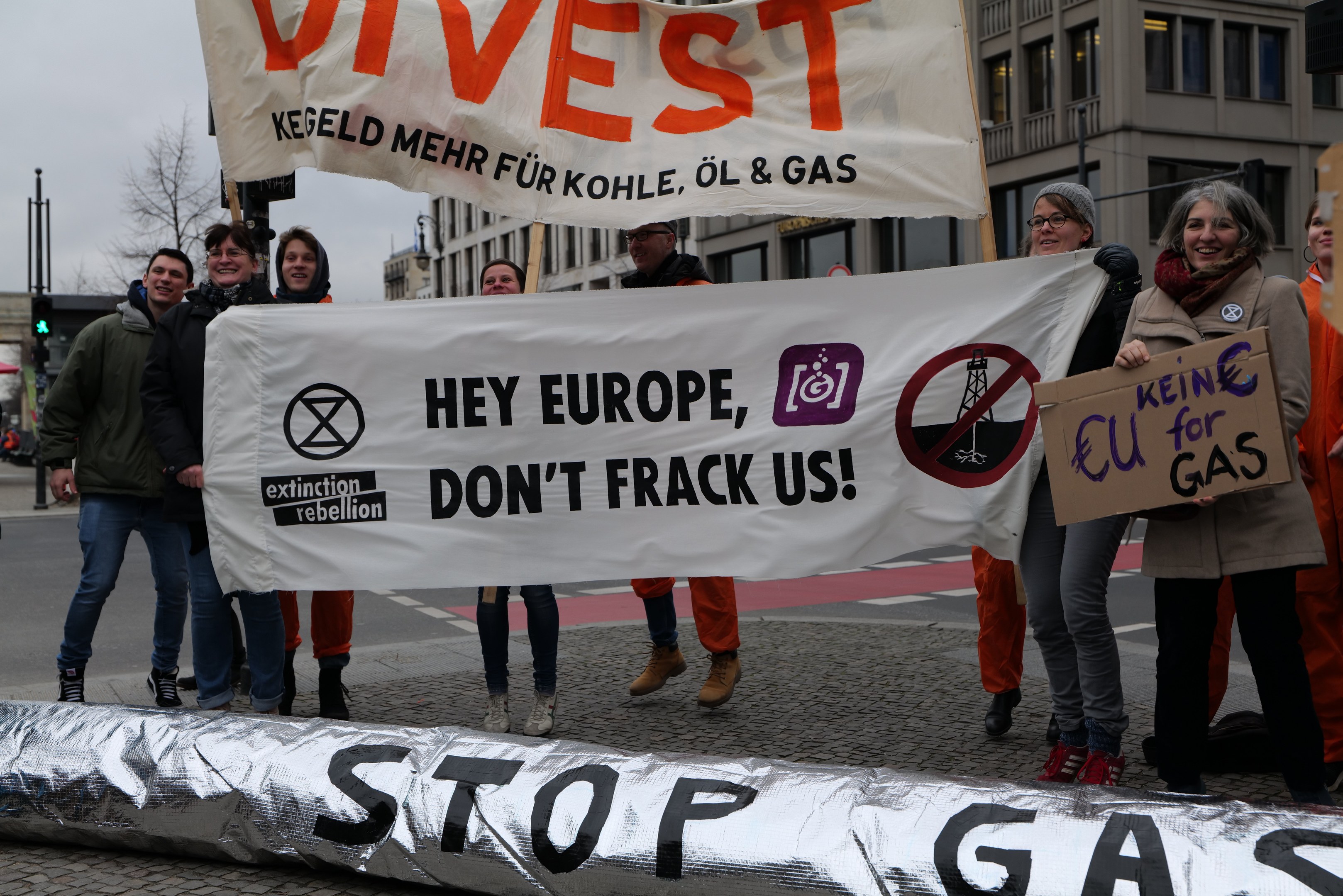 Eine Gruppe von Menschen steht auf einer Straße und hält ein Schild hoch, auf dem "Stoppt Gas" steht, als Protest gegen die Europäische Union. Im Hintergrund sind Gebäude, Bäume, Pfähle, Verkehrszeichen und ein klarer blauer Himmel zu sehen.