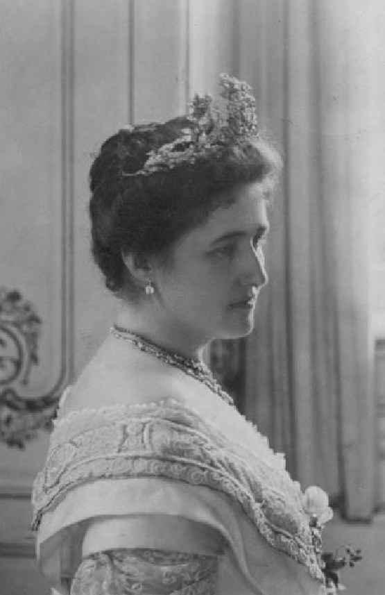 Schwarzes und weißes Foto von Königin Victoria in einem königlichen Kleid, das eine Tiara und eine elegante Hochsteckfrisur trägt und vor einer Vorhangwand steht.
