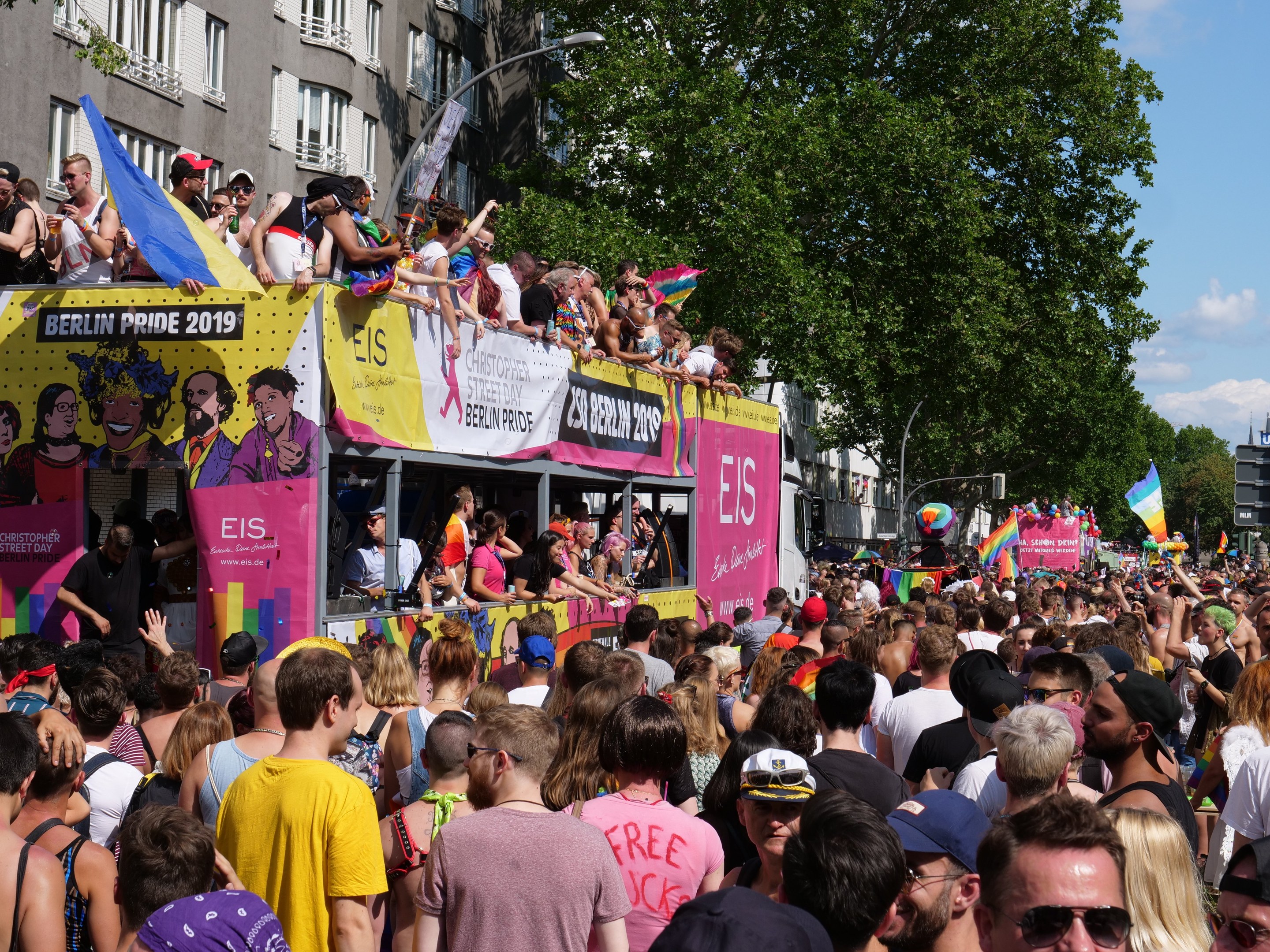 Eine große Menschenmenge marschiert auf der Straße bei der Christopher Street Day Parade 2019 in Berlin, viele tragen Mützen und Schutzbrillen, halten Fahnen und Transparente, mit Gebäuden, Laternenmasten und Bäumen im Hintergrund unter einem bewölkten Himmel.