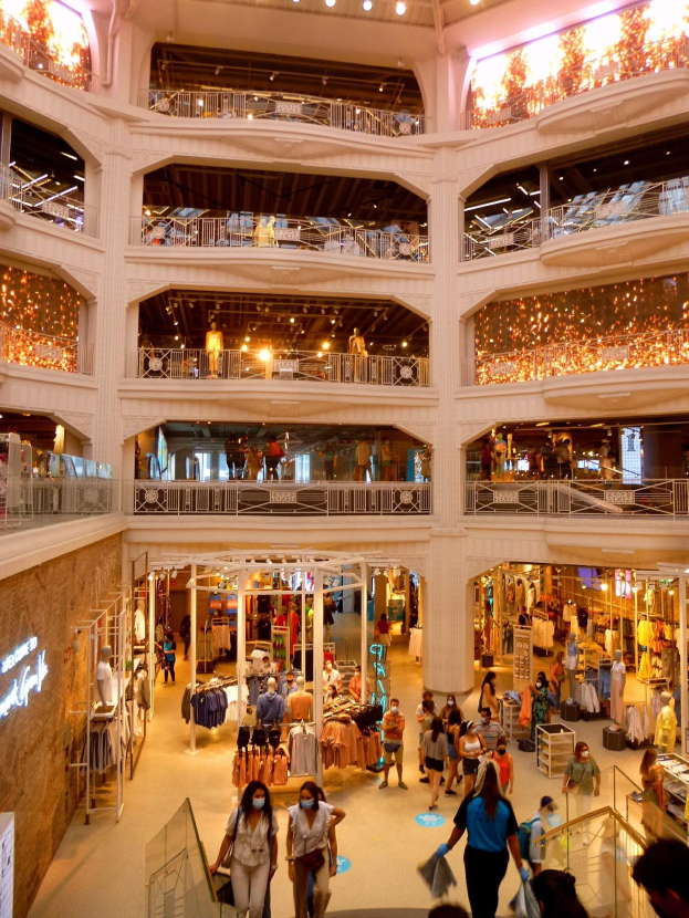 Ein belebtes Einkaufszentrum mit Menschen, die Taschen tragen und Schaufensterpuppen in Kleidern umgeben von Geländern, Lampen und Wandtafeln.