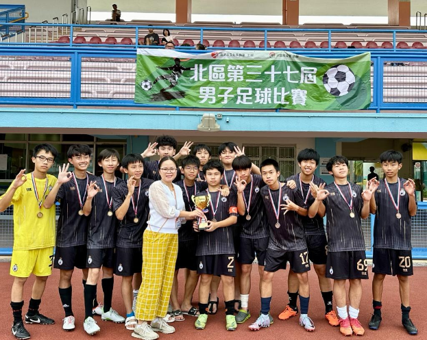 Gruppe junger Männer in Fußballtrikots auf einem Feld stehend, mit Medaillen und einem Pokal haltend, mit einem 'Yokohama U-16 Boys Soccer Team'-Banner im Hintergrund und sitzenden Zuschauern.