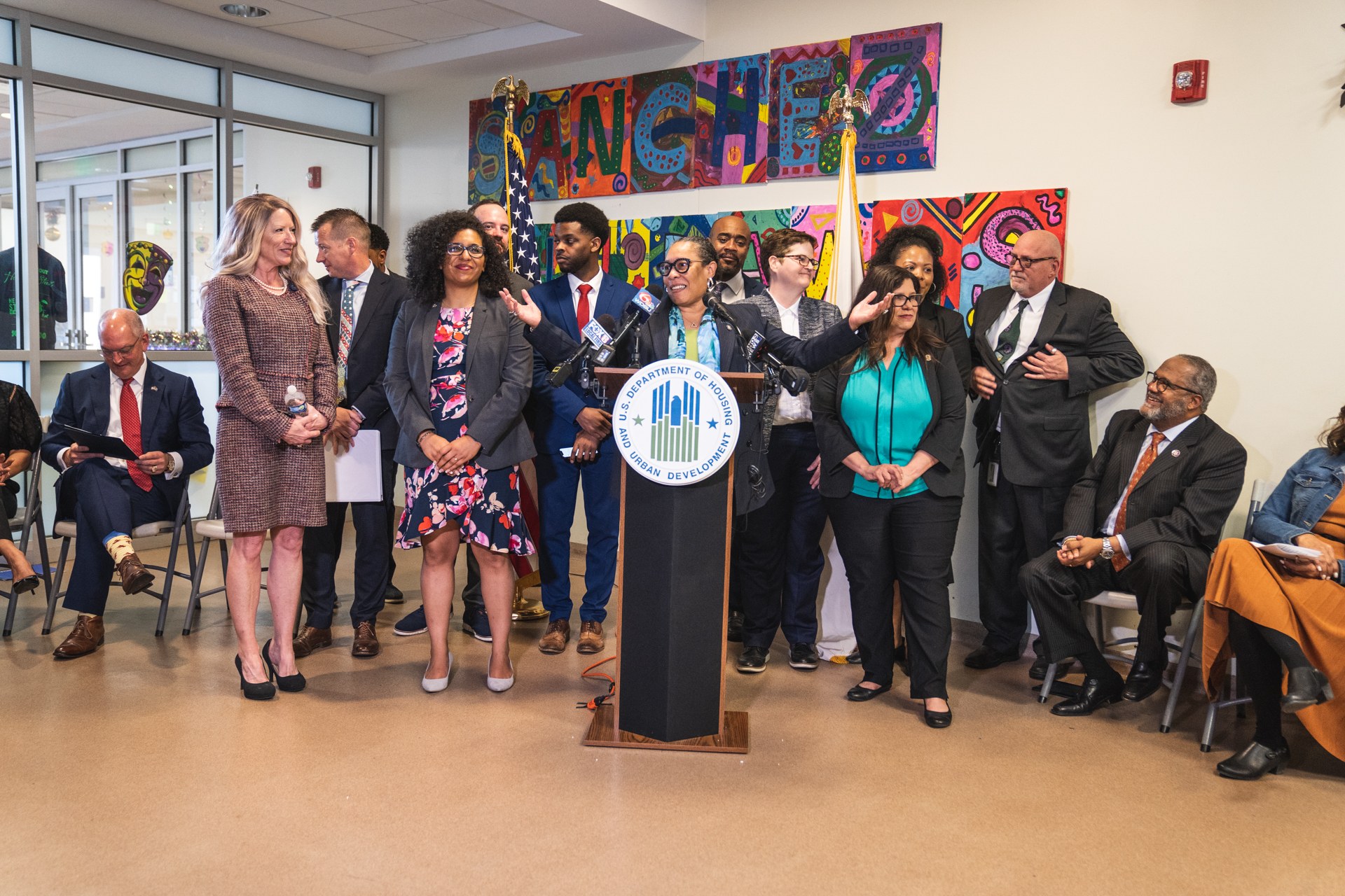 A group of people stands in front of a podium with a microphone and a board displaying text, while others sit on chairs or stand nearby; flags, a fire extinguisher, and ceiling lights are visible on the back wall.