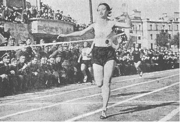 Schwarz-weißes Foto einer Frau bei der 100m-Final der Frauen bei den Olympischen Spielen 1956 auf einer Laufbahn, mit Zuschauern, Gebäuden und Stangen im Hintergrund.
