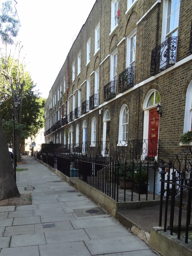 Eine Reihe von Reihenhäusern mit Fenstern, Türen, Geländern, Topfpflanzen, einem Metallzaun, einem Straßenpfosten, Bäumen und einem bewölkten Himmel in London.