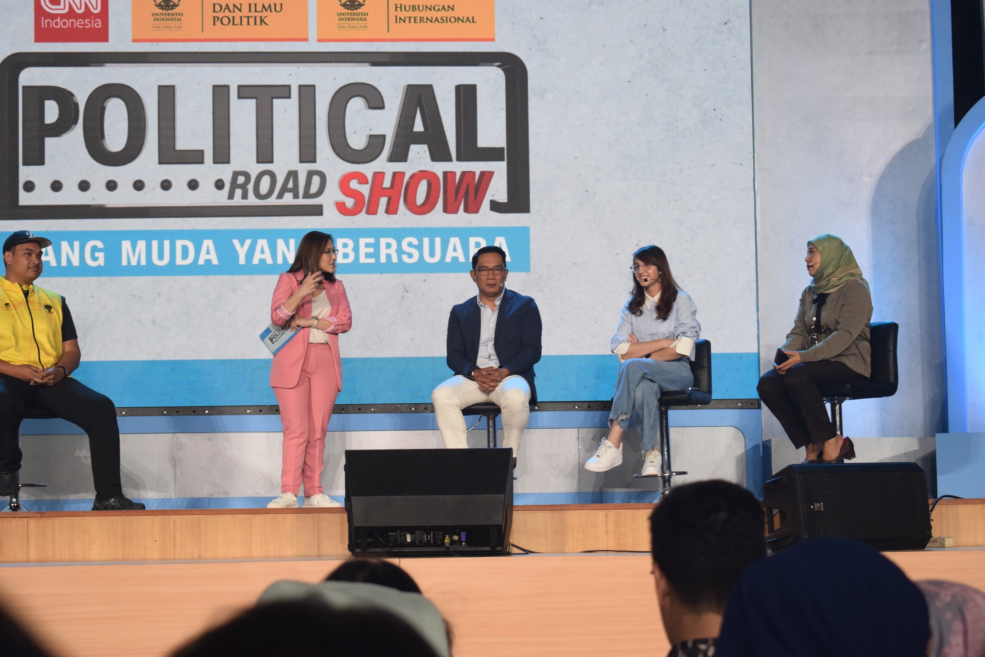 Eine Frau mit einem Mikrofon spricht zu einem Publikum auf einer Bähne, mit den Käpfen der Zuhörer unten und einem 'Political Road Show 2019'-Plakat im Hintergrund.